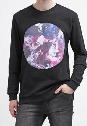 Person trägt ein schwarzes Langarmshirt mit einem großen kreisförmigen Motiv aus bunten violetten, pinken und weißen Rauchschwaden auf der Brust.