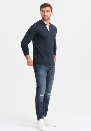 Hombre vistiendo una camiseta henley de manga larga en color marino, jeans azules desgastados y zapatillas blancas, de pie con las manos cerca de los bolsillos contra un fondo blanco.