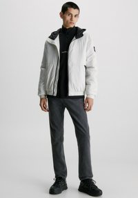 Veste blanche matelassée avec capuche noire, présentant une fermeture éclair et des poignets côtelés. Portée sur un haut noir à col montant et un jean gris. Chaussures noires.