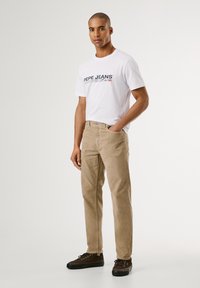 Pantaloni in corduroy beige, t-shirt bianca a girocollo con logo e scarpe marroni con lacci. Il modello è in piedi con le mani in tasca su uno sfondo semplice.