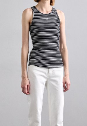 Person, die ein eng anliegendes schwarz-weißes Tanktop mit horizontalen Streifen trägt, kombiniert mit weißen, hoch taillierten Hosen, stehend vor einem einfarbigen Hintergrund.