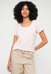 Heled roosa lühikeste varrukatega T-särk ümmarguse kaelusega ja rinnal tagasihoidliku tikandiga logo, kombinatsioonis beežide pükste ja vööga.