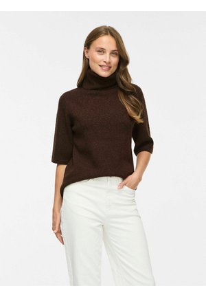 Femme aux longs cheveux châtain clair portant un pull à col roulé marron foncé avec des manches aux coudes et un pantalon blanc, souriante, une main dans la poche.