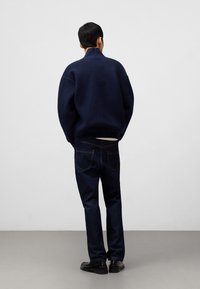 Mörkblå stickad tröja med hög krage, avslappnad passform. I kombination med mörkblå jeans. Modell vänd bort, bär svarta skor. Enkel design.