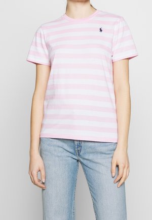 Osoba nosząca jasnoróżowy i biały t-shirt z poziomymi paskami na krótkich rękawach, z małym granatowym logo na lewej piersi oraz jasnoniebieskie dżinsy.