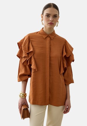 Kvinde iført en rustfarvet bluse med store flæseærmer, cremefarvede bukser, guldbelagte øreringe, armbånd og holder en matchende rustfarvet clutchtaske.