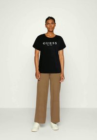 Mulher em pé, vestindo uma camisola preta "GUESS", calças largas castanhas e ténis brancos, contra um fundo claro.
