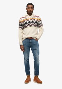 Suéter crema con patrones geométricos multicolores, jeans azules y botas marrones con cordones, con un cuello vuelto.