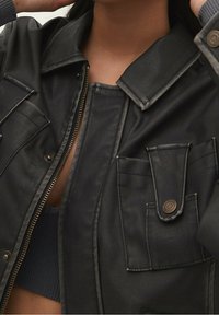 Černá kožená bunda s velkými klopovými kapsami, stříbrným zipem a strukturovaným límcem. Jemná struktura s tlumenými kovovými akcenty.