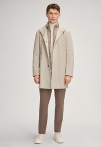 Beige houndstoothjas met een geknipte kraag, gelaagd over een lichtgrijs gewatteerd vest, gecombineerd met bruine broeken en witte sneakers.