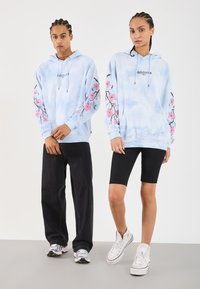 Hoodie azul claro com padrão tie-dye, apresentando designs florais em rosa nas mangas, combinado com calções pretos e sapatilhas brancas. Ajuste casual unissexo.