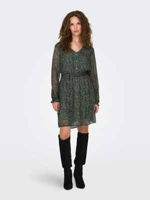 Donna con capelli ricci che indossa un vestito a maniche lunghe verde scuro con motivo e stivali neri in camoscio al ginocchio, in piedi davanti a uno sfondo semplice.