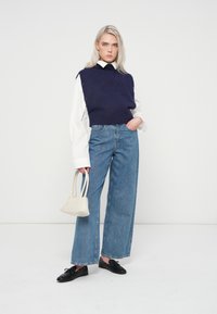 Colete de malha azul-marinho sobre uma camisa branca, combinado com calças largas de denim azul de cintura alta. Sapatos pretos e uma pequena mala bege completam o look.