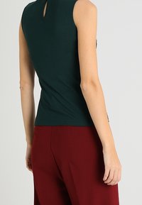 Top verde bosque sin mangas con un escote tipo ojal en la espalda, combinado con pantalones burdeos. La tela parece suave y ajustada.