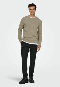 Šedý pletený svetr s kulatým výstřihem, přes který je navlečený bílý košile, doplněný černými slim-fit kalhotami a černými šněrovacími botami.