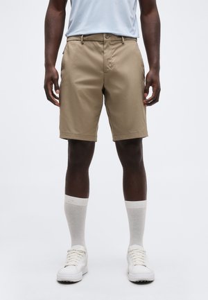 Férfi bézs térdig érő rövidnadrágban, fehér boka zokniban, fehér sportcipőben és egy belezárt világosszürke ingben, egyszínű háttér előtt állva.