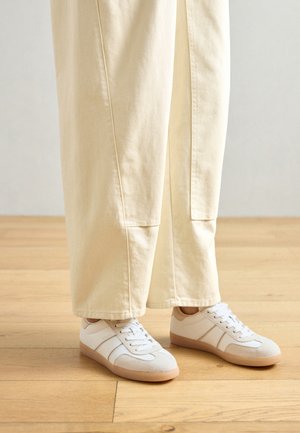 Baskets blanches avec un mélange de textures lisses et en daim, avec des accents beiges et une semelle en caoutchouc, associées à un pantalon large de couleur claire.