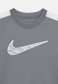 T-shirt de sport gris avec un col rond, arborant un logo Nike blanc avec des détails rayés sur le devant. Le tissu présente une texture lisse.
