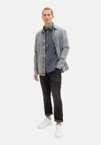Ljus denimjacka över en mörk chambray-skjorta, kombinerat med svarta jeans och vita sneakers. Släta texturer och klassisk passform genomgående.