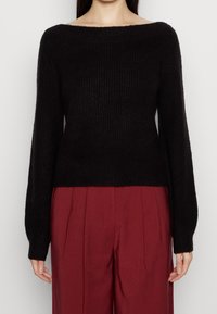 Czarny prążkowany sweter z szerokim dekoltem typu łódka, długimi rękawami i miękką teksturą. Noszony z czerwonymi spodniami o wysokim stanie.