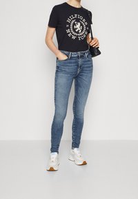 Marinblå t-shirt med vitt grafiskt logo, i kombination med slim fit blå denimjeans. Vita sneakers med bruna detaljer fulländar looken.