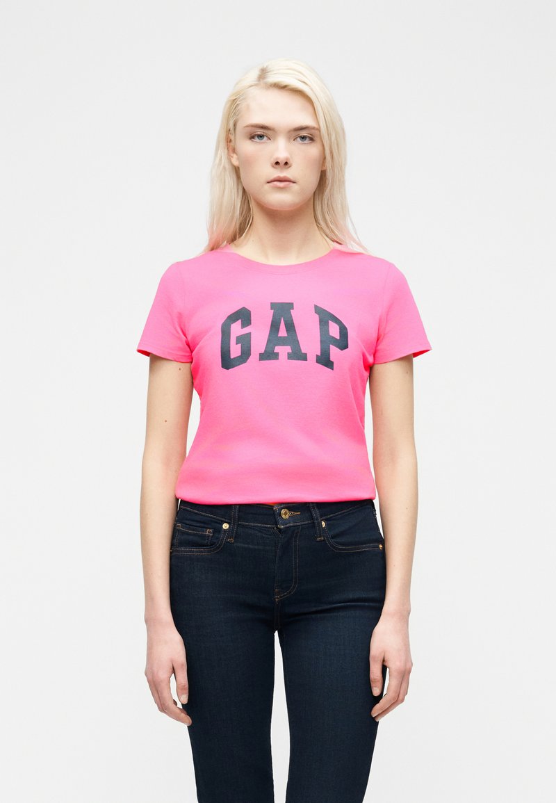 Νεαρή γυναίκα με μακριά ξανθά μαλλιά, ντυμένη με φωτεινή ροζ μπλούζα GAP και σκούρο μπλε τζιν, στέκεται μπροστά σε απλό λευκό φόντο.