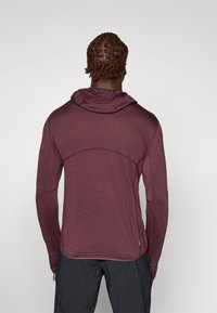 Bordeaux farvet langærmet hættebluse med fin tekstur, buede sømme og tætsiddende design. Komplementerer sorte bukser med lynlåslommer.