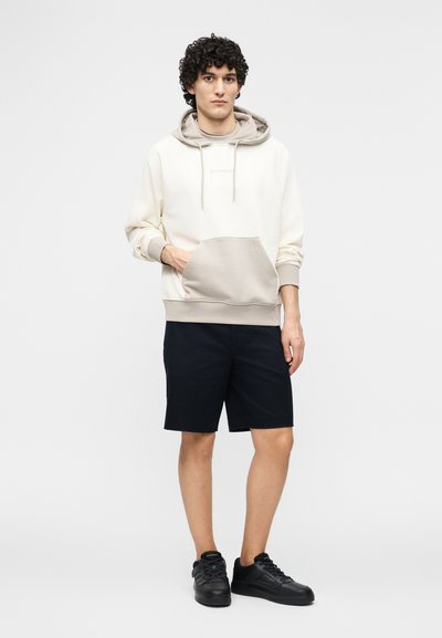 Νεαρός με κατσαρά μαλλιά που φοράει μια εκρού και γκρι μπλούζα με κουκούλα, μαύρο σορτς και μαύρα αθλητικά παπούτσια, στέκεται μπροστά σε απλό φόντο.