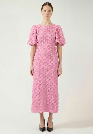 Kvinde iført en lyserød tekstureret midikjole med puffede ærmer og sorte spidse hæle, stående foran en ensartet, lys baggrund.