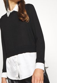 Donna che indossa un maglione corto nero a strati sopra una camicia bianca con colletto e maniche lunghe, insieme a pantaloni in pelle nera lucida.