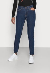 Blaue Denim-Jeans mit schlanker Passform, hoher Taille, fünf Taschen und einem kleinen Messingknopf. Getragen mit weißen Sneakers und einem schwarzen Langarmoberteil.