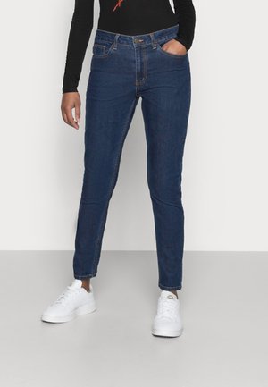 Blauwe denim jeans met een slim fit, hoge taille, vijf zakken en een kleine messing knoop. Draagt een wit sneakers en een zwart langemouwen shirt.