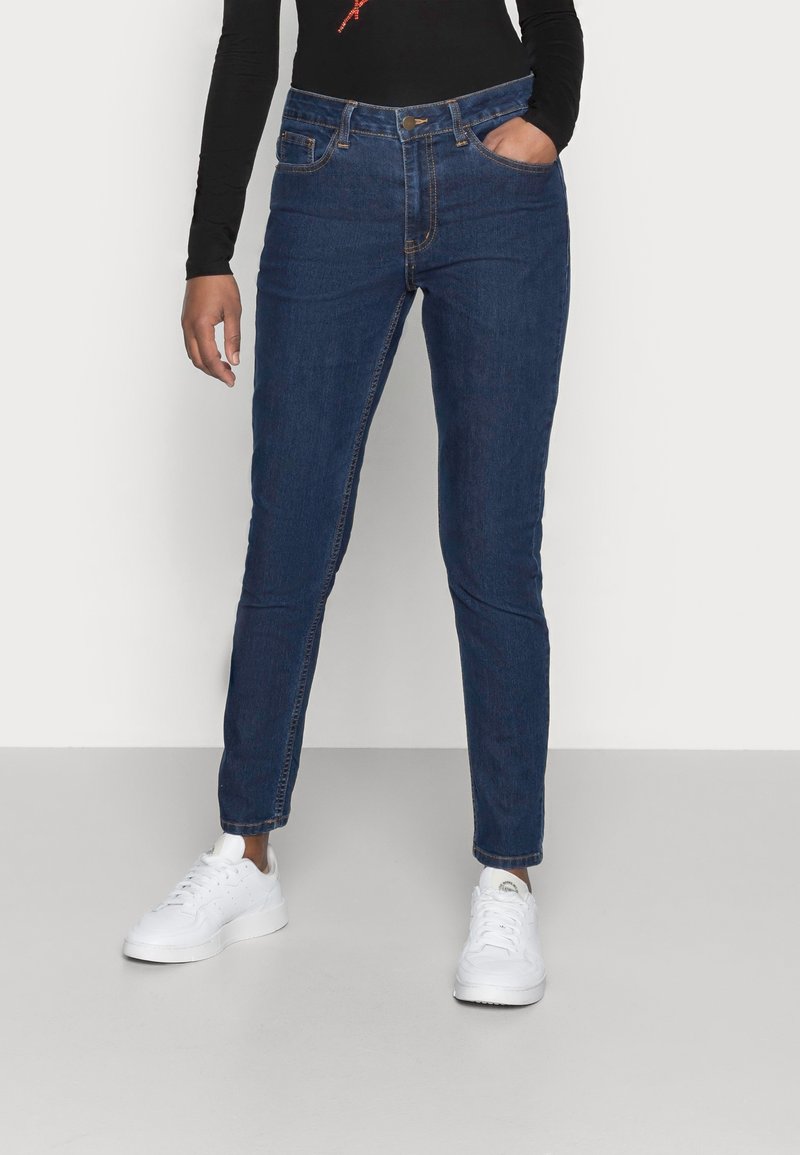 Blaue Denim-Jeans mit schlanker Passform, hoher Taille, fünf Taschen und einem kleinen Messingknopf. Getragen mit weißen Sneakers und einem schwarzen Langarmoberteil.