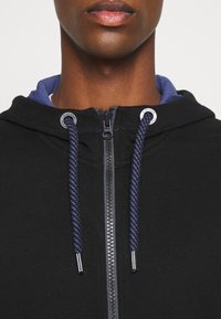 Sweat à capuche noir avec fermeture éclair et doublure intérieure bleu marine ; doté de cordons épais bleus et d'accents en métal argenté.