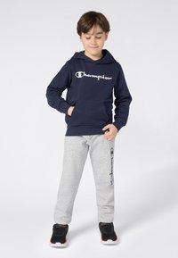 Jongentje in een marineblauwe hoodie en grijze joggingsbroek met logo's, staande met één hand in zijn zak en kijkend naar beneden, op een effen lichte achtergrond.