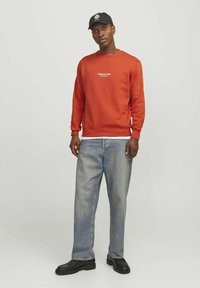 Rode sweatshirt met geribbelde boorden en zoom, witte logo op de borst, gecombineerd met lichtblauwe denim jeans en zwarte casual schoenen. Zwarte pet gedragen.