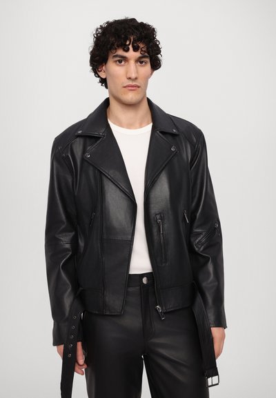Zwarte leren motorjas met ritszakken, drukknopenkraag en verstelbare riem. Draag het over een wit geribbeld shirt en zwarte broek.