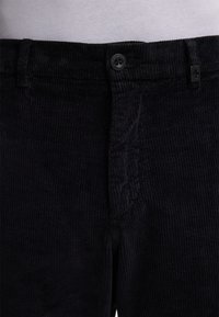 Black corduroy pants featuring a button closure, vertical ribbed texture, and styled with tailored seams and a front pocket detail.