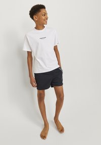 Camiseta blanca de algodón con logotipo de texto negro, combinada con pantalones cortos oscuros de navy con detalles gráficos blancos. El modelo está descalzo.