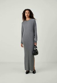 Γκρι ραβδωτό πλεκτό μακρύ φόρεμα με στρογγυλή λαιμόκοψη, πλαινές σχισμές και μακ�ριά μανίκια, συνδυασμένο με μια μικρή μαύρη τσάντα, σε ανοιχτό φόντο.