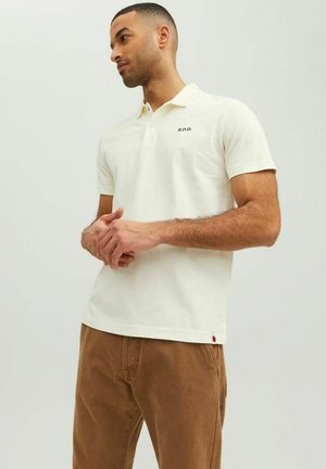 Polo shirt en coton crème à manches courtes, avec un col à trois boutons et un petit "R.D.D." brodé sur la poitrine. Pantalon marron.