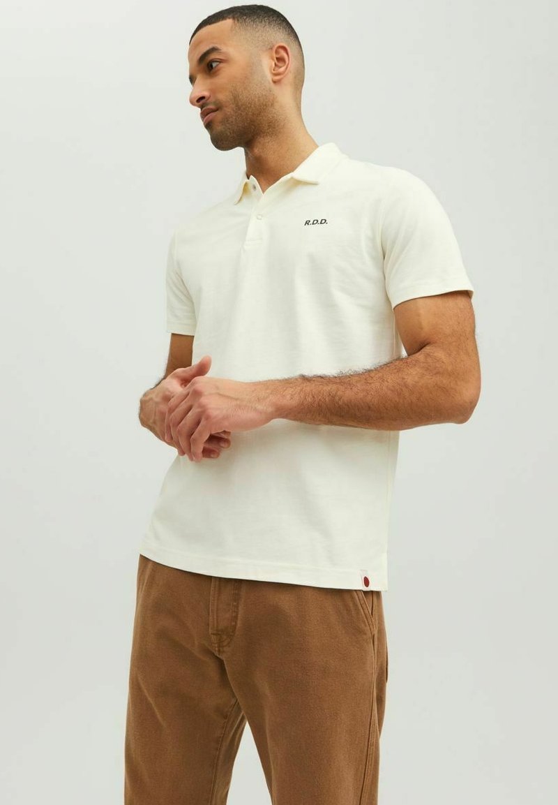 Polo shirt en coton crème à manches courtes, avec un col à trois boutons et un petit "R.D.D." brodé sur la poitrine. Pantalon marron.