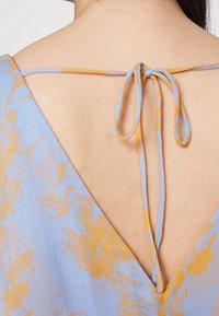 Vestido floral azul-claro com costas em forma de V e fechamento por laço. O tecido é transparente com uma textura suave e detalhes amarelos no padrão.