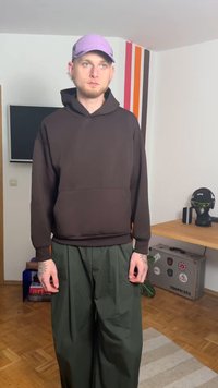 Felpa marrone con tasca frontale, abbinata a pantaloni verdi larghi. Modello con cappello viola chiaro. Ambiente interno con arredamento minimalista.