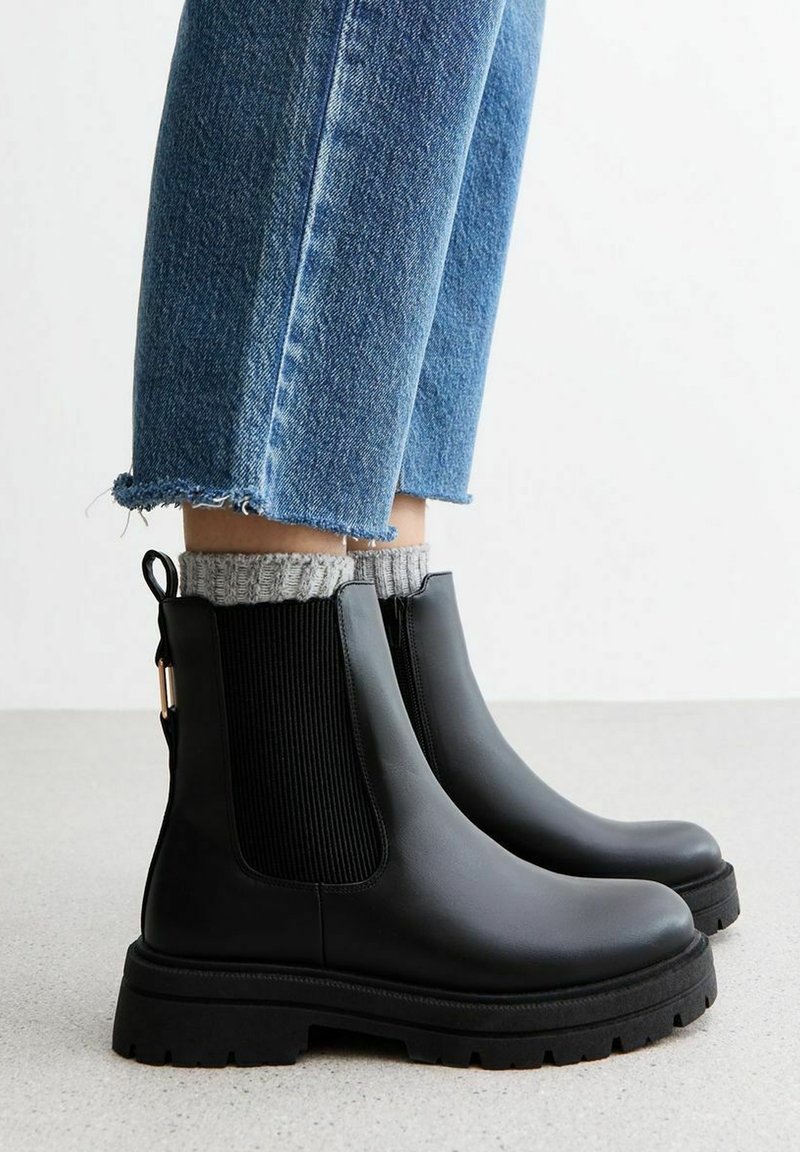 Bottes Chelsea en cuir noir avec panneaux élastiques côtelés sur les côtés et semelle en caoutchouc texturée, associées à des chaussettes grises en maille et un denim court.
