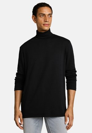 Man wearing black long-sleeve turtleneck shirt and light gray pants, standing against plain light background, looking slightly right.