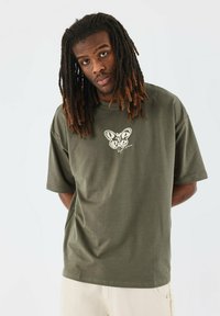 Camiseta de algodón verde oliva con mangas cortas, que presenta un gráfico de mariposa blanca en el pecho. Diseño de ajuste relajado y cuello redondo.