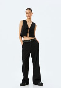 Schwarze Crop-Weste mit tiefem V-Ausschnitt und zwei vorderen Bindebändern, kombiniert mit hochtaillierten schwarzen Hosen und klobigen schwarzen Schuhen.
