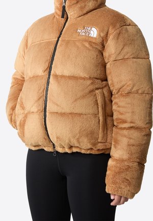 Doudoune en peluche marron clair avec fermeture éclair noire et logo The North Face blanc sur la poitrine, portée avec un pantalon noir.