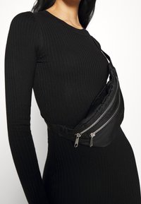 Marsupio nero in tessuto con due tasche con cerniera argentata e un design elegante e curvo, indossato attraversando il torso sopra un vestito lungo a maniche lunghe nero a coste.
